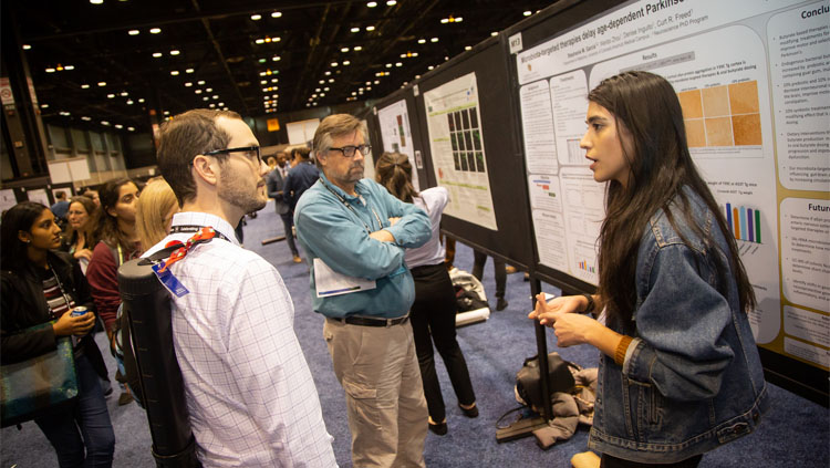 Two men listening to a woman explain her poster presentation at Neuroscience 2019.