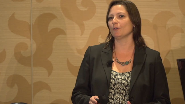 Victoria Puig stands in front of a tan wall wearing a black jacket while giving a lecture.