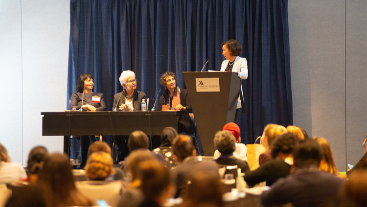 Three panelists sit at a table while a moderator stands at a podium at the celebration of women in neuroscience luncheon 2019