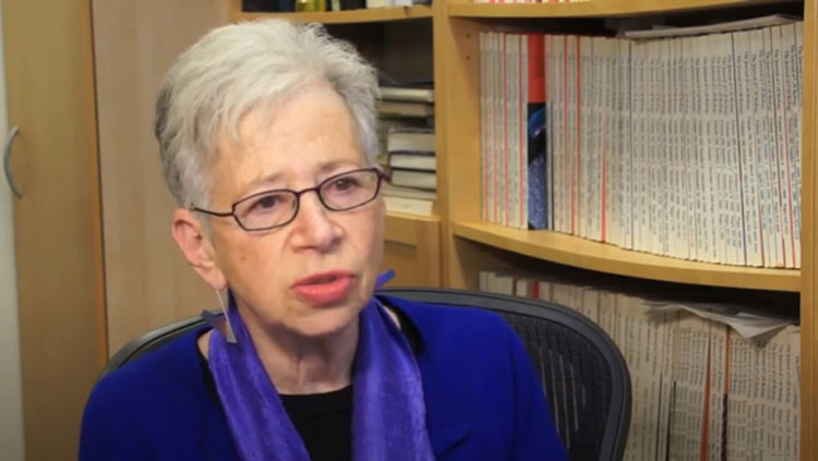 A woman wearing a blue shirt and glasses speaks to the camera with a bookshelf behind her.
