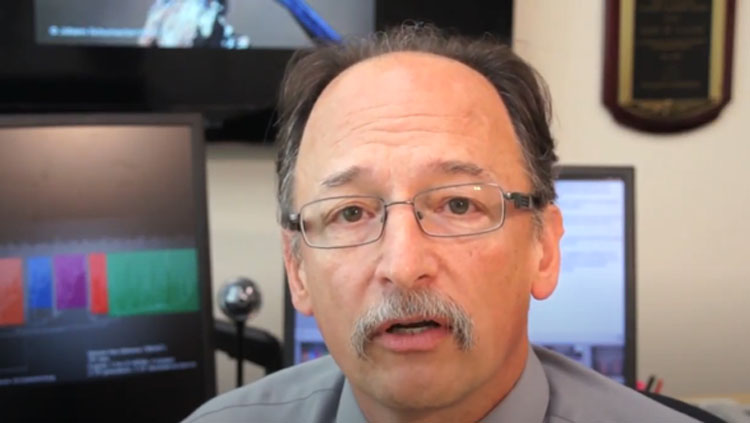 A man with a mustache and glasses speaks to a camera.