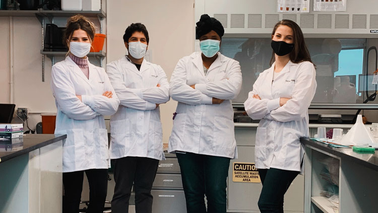 Four scientists stand in front of their lab benches wearing lab coats with their arms crossed.