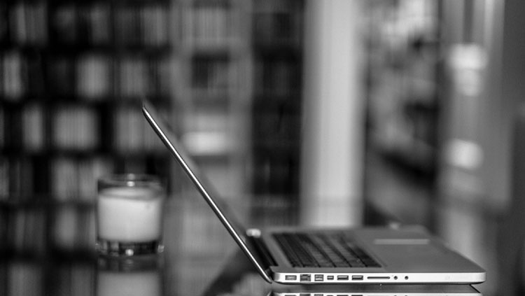 Side view of a laptop sitting open on glass table with a bookshelf just out of focus in the background.