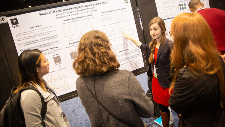 Three people listen as a young woman explains her research poster at Neuroscience 2019. 