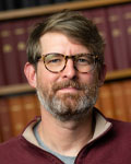 Headshot of Timothy Denison in front of a book shelf wearing glasses and a red sweater.